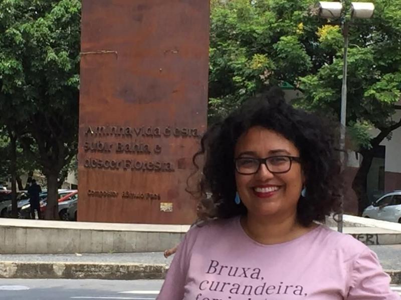 A imagem mostra uma mulher sorridente de cabelos cacheados e óculos, vestindo uma camiseta rosa com os dizeres "Bruxa, curandeira." Ela está em um espaço urbano com árvores ao fundo e uma placa metálica enferrujada com uma inscrição em relevo. A frase na placa inclui "A minha vida é subir Bahia e descer Floresta", uma referência à cidade de Salvador, BA. O ambiente sugere um local público, possivelmente um ponto turístico ou cultural.