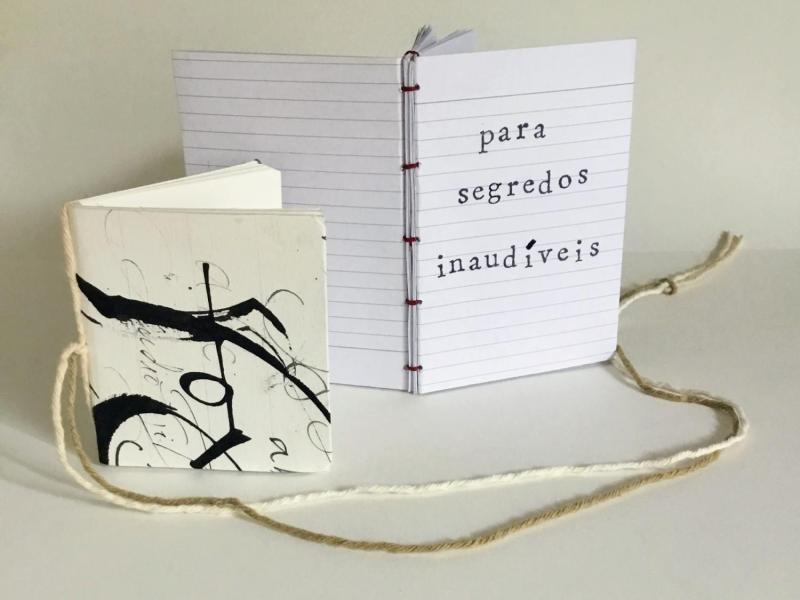 A imagem apresenta dois cadernos artesanais sobre uma superfície branca. O caderno à direita está aberto, revelando páginas pautadas e a frase “para segredos inaudíveis” escrita em letra mecânica. Sua encadernação é feita com costura exposta em fio vermelho. O caderno à esquerda é menor, tem capa decorada com traços caligráficos abstratos em preto sobre fundo branco e está fechado. Um barbante cru e uma linha vermelha conectam os cadernos, sugerindo uma relação entre eles, remetendo a um conceito artístico.