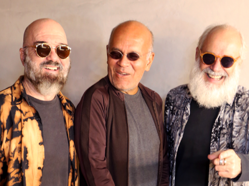 A imagem mostra os músicos Carlos Malta, Marcos Suzano e Alex Meirelles sorrindo em um clima descontraído. Todos usam óculos escuros e exibem visuais marcantes. Um deles tem barba cheia e veste uma camisa estampada em tons de preto e laranja, outro usa uma jaqueta marrom sobre uma camiseta cinza, e o terceiro tem uma longa barba branca e veste uma camisa estampada em preto e branco. A cena transmite cumplicidade e energia, refletindo a sintonia entre os artistas.