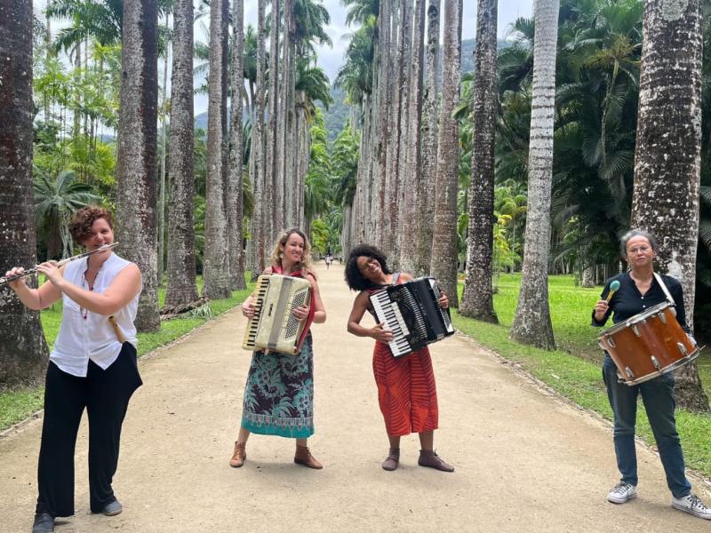 A imagem mostra as integrantes do grupo de forró Luz de Candeeiro em uma alameda cercada por palmeiras imperiais. Da esquerda para a direita, estão: Maria Souto tocando flauta, Marcela Coelho e Joana Araújo nas sanfonas, e Georgia Camara na percussão. As musicistas estão sorrindo e transmitindo alegria em meio ao cenário tropical, unindo natureza e cultura popular em uma atmosfera leve e vibrante.