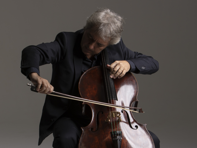 O violoncelista Hugo Pilger aparece em um ambiente neutro, com fundo cinza e iluminação suave. Vestido com roupa escura, ele está concentrado enquanto toca seu violoncelo, inclinando levemente o corpo para a frente e movimentando o arco com precisão. A pose transmite intensidade e domínio técnico, refletindo a expressividade e o refinamento de sua performance musical.