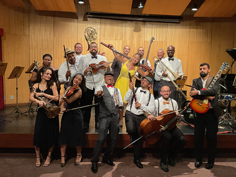 A imagem mostra um grupo musical posando de forma descontraída em um palco com parede de madeira e instrumentos ao fundo. São quatorze integrantes, todos sorridentes e vestidos elegantemente — alguns com trajes formais, como gravatas borboleta, e outros com roupas mais coloridas. Entre os instrumentos estão violinos, violoncelo, guitarra, contrabaixo, sopros e percussão. A atmosfera é alegre e celebrativa, transmitindo o entusiasmo coletivo pela música.