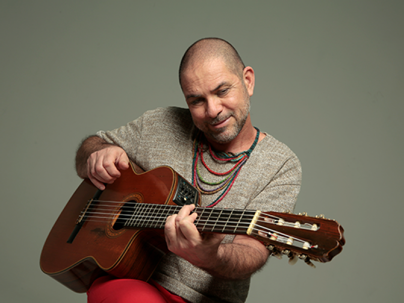 A imagem mostra o cantor e compositor paulistano Paulo Padilha tocando violão. Ele está sorrindo levemente, em uma pose descontraída, usando uma camiseta cinza, calça vermelha e colares coloridos. O fundo é neutro, destacando Padilha e seu instrumento. A cena transmite leveza e intimidade, refletindo o estilo bem-humorado e envolvente do artista.