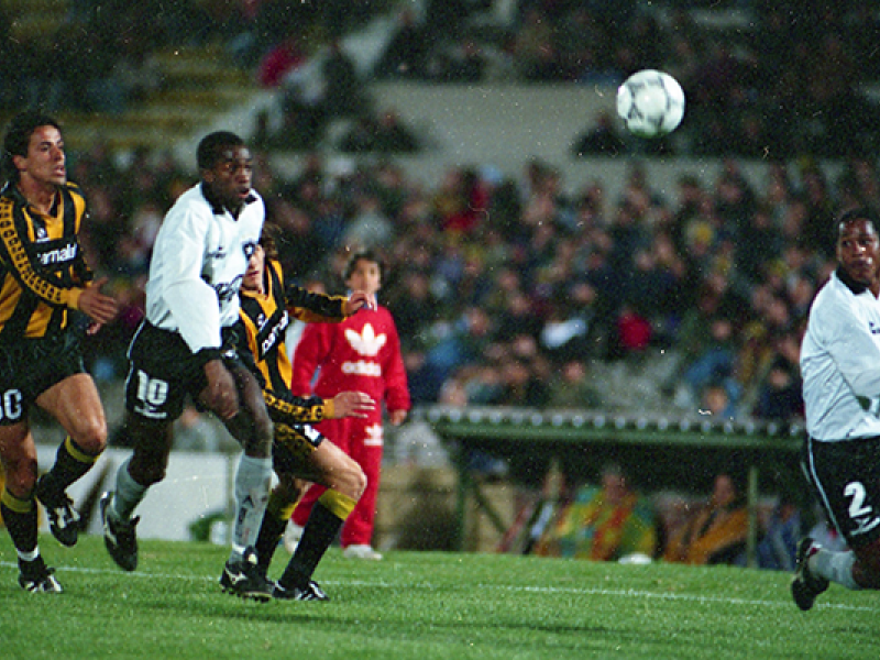 A imagem mostra um momento de intensa disputa durante uma partida entre Peñarol e Botafogo. Jogadores do Peñarol, com uniforme listrado em preto e amarelo, e do Botafogo, com uniforme branco e preto, acompanham atentos a trajetória da bola no ar. A cena se passa em um estádio com arquibancadas lotadas, transmitindo a energia vibrante de um jogo competitivo. Ao fundo, um garoto com roupa adidas vermelha observa a ação de perto.