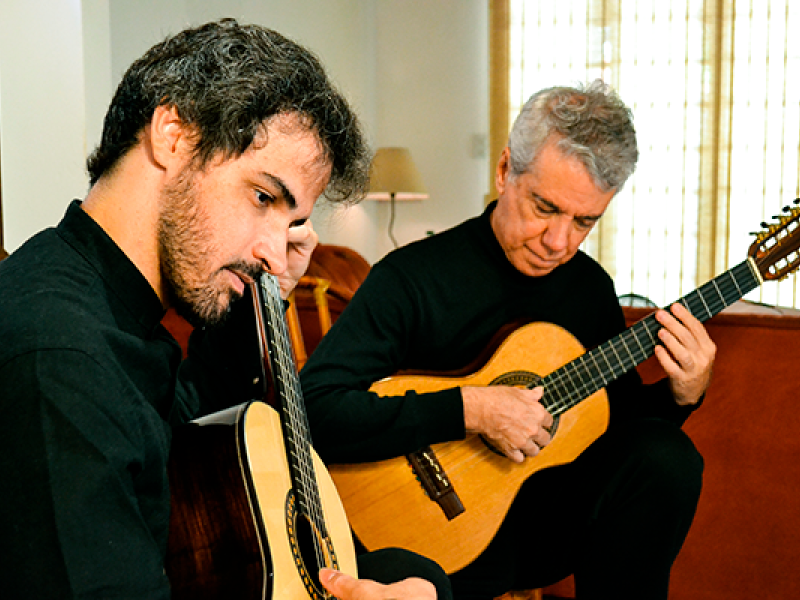 Os músicos que formam o Duo Roberto Victório e Sergio Ribeiro, estão sentados em um ambiente interno, tocando violões com concentração e sensibilidade. Ambos vestem roupas escuras. Um deles tem barba e cabelo escuro e cacheado; o outro tem cabelo grisalho. A cena transmite um momento íntimo de execução musical, com luz natural entrando por uma janela ao fundo e criando uma atmosfera acolhedora.
