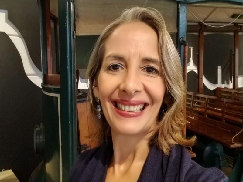 A escritora e artista plástica Patrícia Montês sorri para a câmera em um ambiente que remete a um espaço expositivo ou museu. Ela usa um casaco azul escuro e brincos delicados, e seus cabelos lisos e claros têm mechas grisalhas. Ao fundo, vê-se parte de um antigo veículo com bancos de madeira — possivelmente um bonde —, sugerindo um contexto cultural ou histórico. A imagem transmite simpatia e envolvimento com o ambiente ao redor.