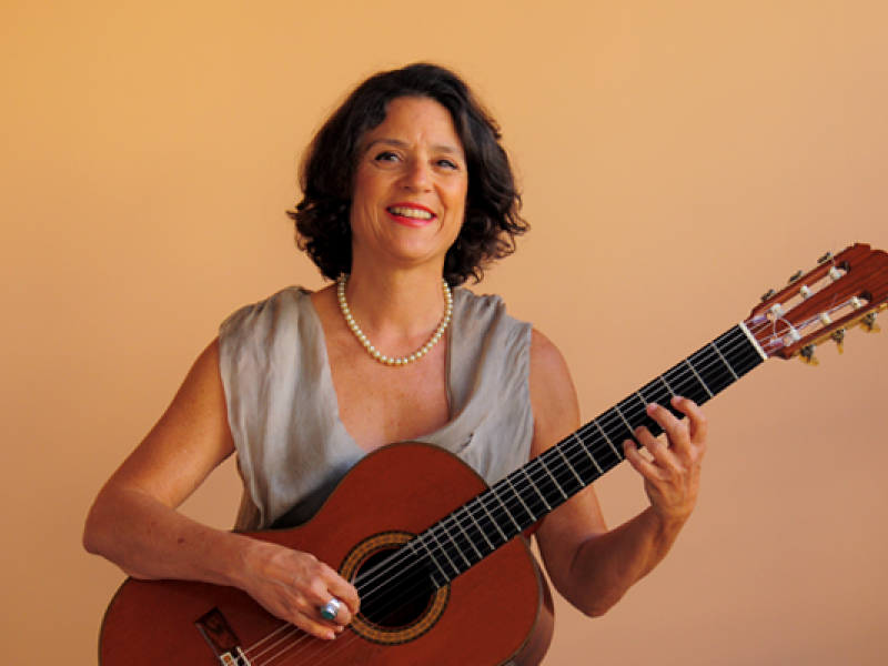 A violonista Adriana Ballesté aparece sorridente, segurando um violão em posição de toque diante de um fundo em tom pêssego. Usa um vestido claro de tecido leve e colar de pérolas, com os cabelos escuros e ondulados na altura dos ombros. A imagem transmite leveza, elegância e afinidade com a música, refletindo uma atmosfera acolhedora e serena.