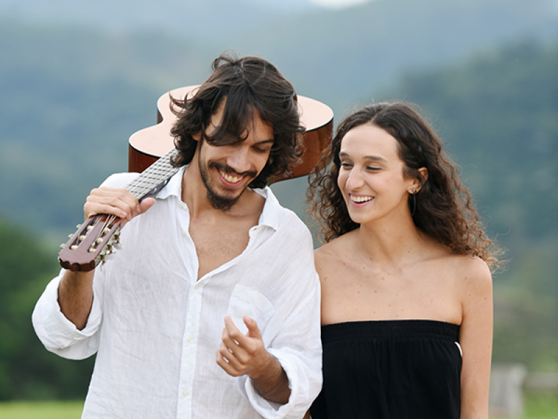Os cantores e violonistas Antonia Medeiros e Guilherme Imia, integrantes do Duo Dois a Dois, caminham ao ar livre em meio à natureza, com montanhas ao fundo. Guilherme, de barba e cabelos ondulados, veste camisa branca e carrega um violão apoiado no ombro. Antonia, de cabelos cacheados e soltos, usa um macacão preto sem alças. Eles trocam sorrisos e parecem conversar, transmitindo leveza, sintonia e conexão artística.