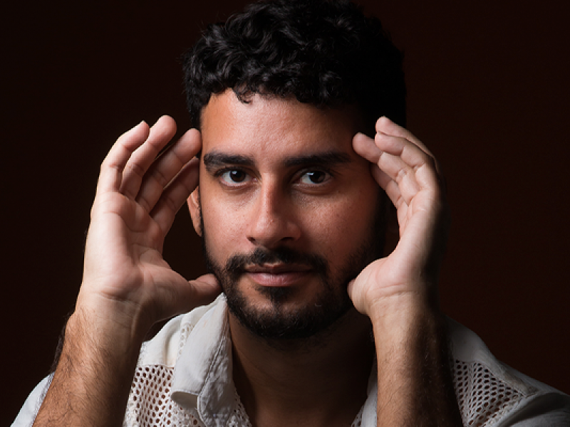 Homem de pele morena, cabelo cacheado curto e barba bem aparada, olha diretamente para a câmera com expressão serena. Ele está com as mãos levantadas próximas ao rosto, como se estivesse enquadrando ou refletindo. Usa uma camisa clara com textura vazada nos ombros. O fundo é escuro, destacando seu rosto iluminado.
