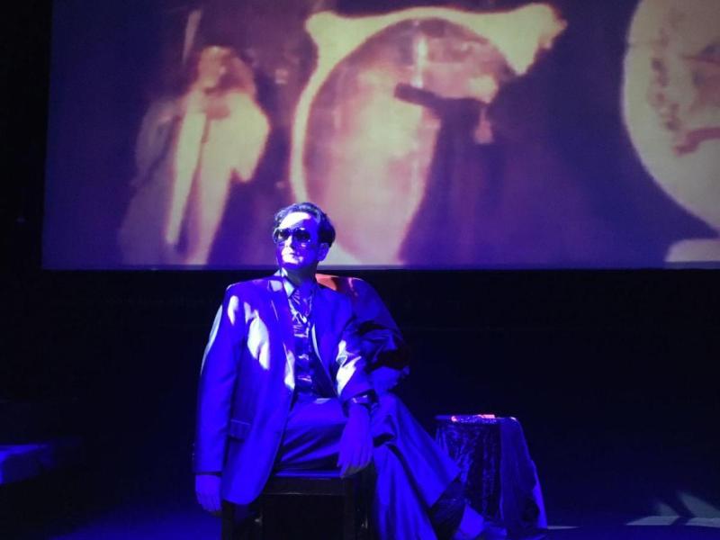Na imagem, uma pessoa sentada em um banco no centro do palco é iluminada por uma luz azul intensa, que cria um efeito dramático. A figura veste um terno e usa óculos escuros, com postura relaxada e expressiva. Ao fundo, uma projeção com imagens difusas e simbólicas contribui para uma atmosfera cinematográfica e enigmática. A composição remete ao universo do teatro performático ou do cinema experimental, evocando mistério e intensidade visual.