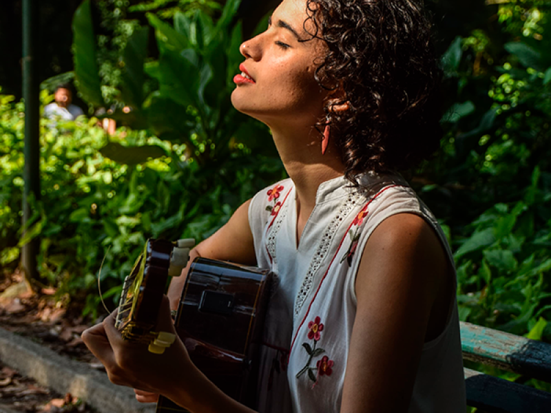 A cantora e violonista Katarina Assef aparece sentada em um banco de madeira em meio à vegetação exuberante, tocando violão. Com cabelos cacheados curtos, ela usa uma roupa clara com detalhes florais. De olhos fechados e rosto levemente inclinado para cima, parece absorver a música. A luz natural destaca seu perfil, criando uma atmosfera serena, sensível e contemplativa.