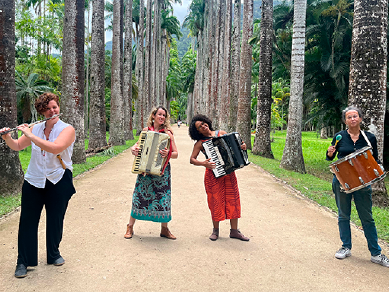 A imagem mostra as integrantes do grupo de forró Luz de Candeeiro em uma alameda cercada por palmeiras imperiais. Da esquerda para a direita, estão: Maria Souto tocando flauta, Marcela Coelho e Joana Araújo nas sanfonas, e Georgia Camara na percussão. As musicistas estão sorrindo e transmitindo alegria em meio ao cenário tropical, unindo natureza e cultura popular em uma atmosfera leve e vibrante.