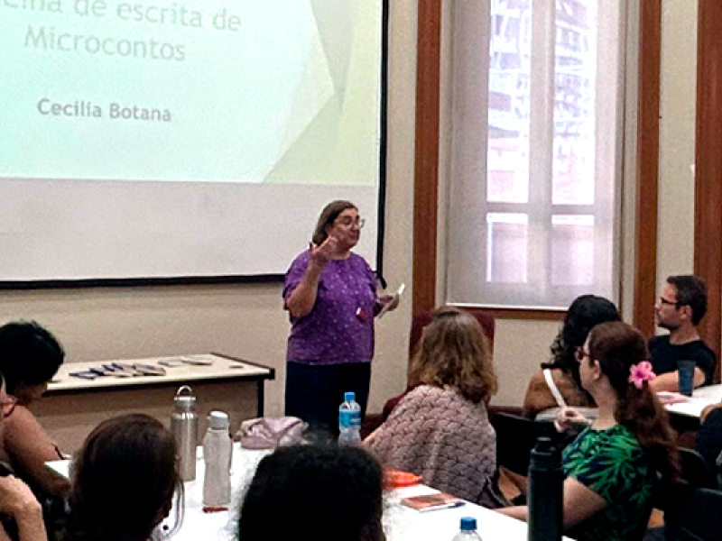 Imagem de oficina de escrita criativa em andamento, conduzida por Cecilia Botana, conforme indicado na projeção ao fundo com o título "Oficina de escrita de Microcontos". Cecilia, veste uma blusa roxa e está em pé diante de uma turma atenta, gesticulando enquanto fala. Os participantes estão sentados em mesas com cadernos e garrafas de água, em um ambiente iluminado por janelas grandes, que indicam tratar-se de uma sala em um prédio antigo, possivelmente histórico. A atmosfera é de concentração e interesse.