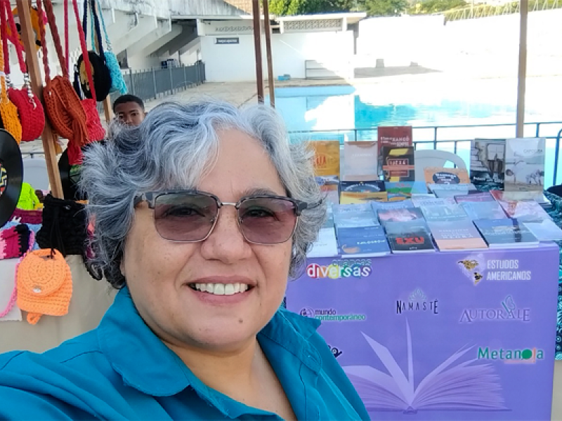 Mulher de cabelos grisalhos e curtos, usando óculos escuros e camisa azul, sorri para a câmera em uma selfie. É Léa Carvalho, publisher da Editora Metanoia, especializada em Gênero, Sexualidade e Diversidade. Ao fundo, há uma mesa com diversos livros expostos sobre uma toalha roxa com palavras como “diversas”, “Metanoia” e “Autor(a)Me”. Também há bolsas coloridas penduradas à esquerda e uma piscina ao fundo, sugerindo um evento ao ar livre, como uma feira cultural ou literária.