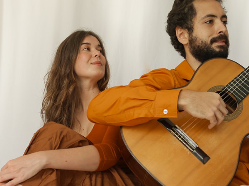Miguel Rabello (voz e violão) e Julia Nali (voz) aparecem sentados lado a lado em um ambiente de luz suave, com fundo claro. Ele toca violão e olha à frente com expressão serena, enquanto ela o observa com um leve sorriso, transmitindo sintonia e cumplicidade. Ambos usam roupas em tons terrosos, que harmonizam com a atmosfera intimista e acolhedora da imagem.