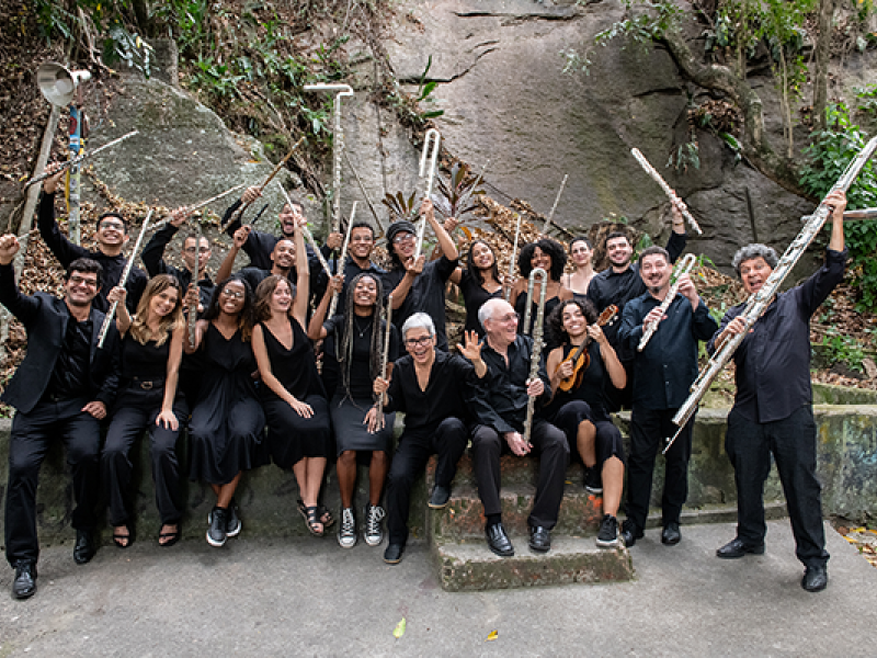 Um grupo grande de músicos posa ao ar livre diante de uma parede de pedra rodeada por vegetação. Todos estão vestidos de preto e exibem seus instrumentos com alegria, como flautas, trombones, violinos e clarinetes. Muitos sorriem, erguendo os instrumentos no ar em um gesto de celebração. A cena transmite um forte senso de união, entusiasmo e espírito coletivo em torno da música.