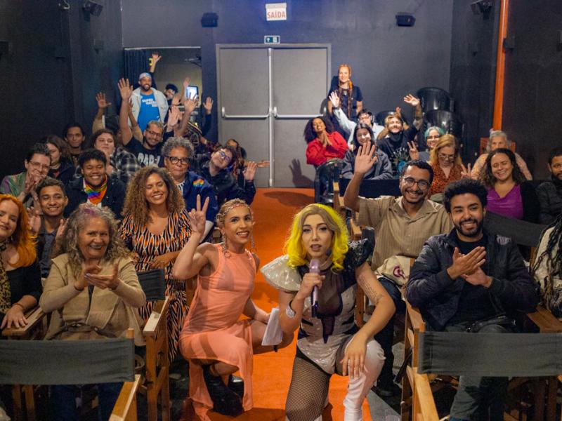 Pessoas de diferentes idades e estilos estão sentadas em um auditório pequeno, sorrindo e acenando para a câmera. Na frente, três pessoas se destacam, incluindo uma drag queen com peruca loira e figurino chamativo, que segura um microfone. O ambiente é acolhedor e descontraído, sugerindo um evento cultural ou artístico com forte senso de comunidade e celebração da diversidade.