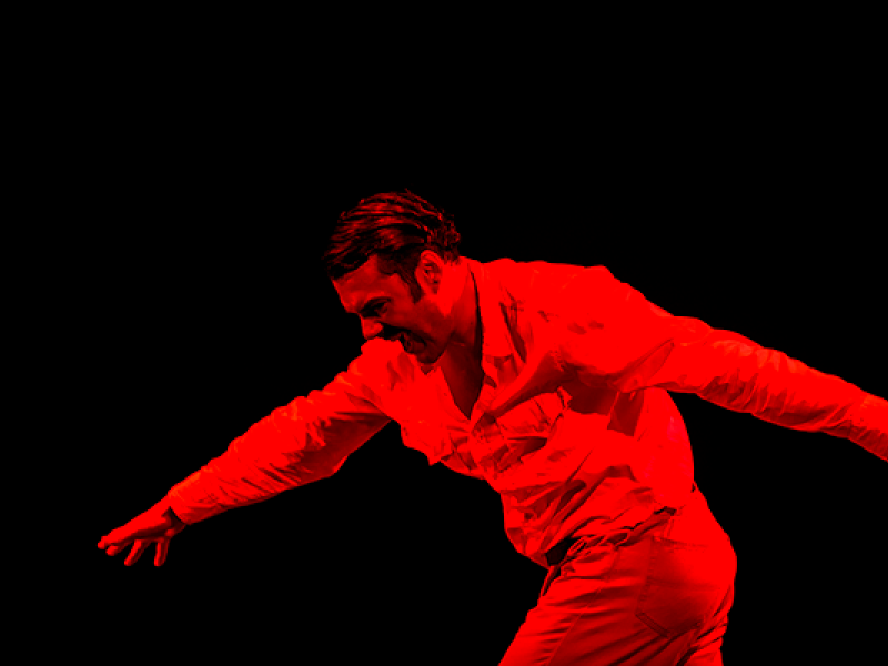 Homem em cena performática com iluminação totalmente vermelha, destacando seu corpo contra o fundo preto. Ele veste roupa social clara e está em movimento, com os braços estendidos e o tronco levemente inclinado para frente, transmitindo intensidade e expressão corporal. A imagem sugere um momento dramático ou coreográfico, com forte carga emocional e estética teatral.