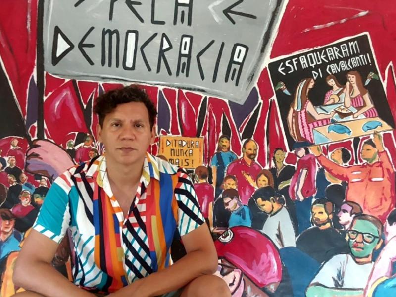 A imagem mostra o artista visual Alex Frechette sentado em frente a um mural vibrante que retrata uma cena de protesto, com cartazes e pessoas reunidas em manifestação. Ele veste uma camisa colorida com listras verticais e tem expressão séria. No fundo, lê-se frases como "Pela Democracia", "Ditadura Nunca Mais!" e "Esfacelaram o Evalantim!".