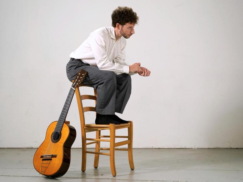 O violonista Giacomo Copiello aparece agachado sobre uma cadeira de madeira, com expressão contemplativa e as mãos entrelaçadas. Veste camisa branca, calça cinza e está descalço. Ao lado da cadeira, um violão clássico está apoiado, sugerindo um momento de introspecção artística ou preparação para uma performance. O ambiente é minimalista, com fundo branco e piso claro, destacando a presença do músico.