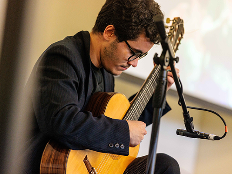 O violonista Tomas Haushahn (RS/EUA) toca com concentração, sentado diante de um microfone. Usa óculos, blazer escuro e camiseta preta. A imagem capta o gesto preciso de sua performance musical, com destaque para o violão e a expressão focada do artista. Ao fundo, uma tela desfocada sugere que ele se apresenta em um evento cultural ou recital.