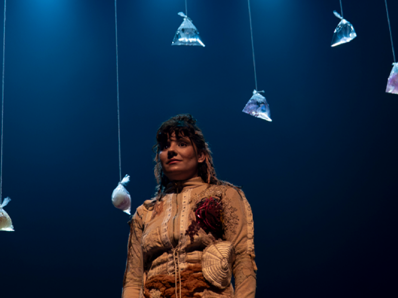 Cena teatral com iluminação azul. Uma mulher está no centro do palco, vestida com um figurino artístico com texturas de conchas e um coração vermelho aparente no peito. Do teto pendem vários saquinhos plásticos com água e pequenos peixes ou elementos marinhos dentro, como os usados para transportar peixes de aquário. A atmosfera é poética e simbólica.