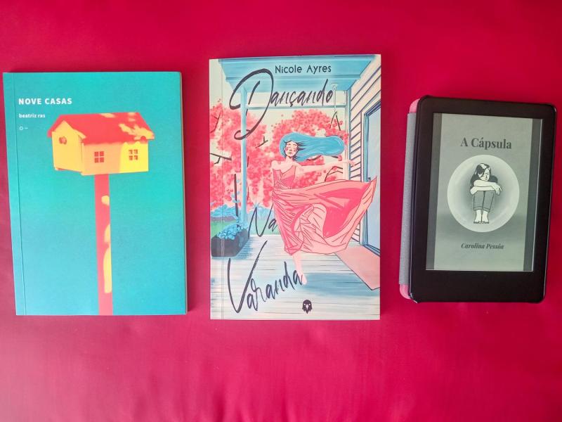 Imagem mostra três livros dispostos lado a lado sobre uma superfície vermelha. À esquerda está "Nove Casas", de Beatriz Pizani, com capa azul e a imagem de uma casa amarela sobre uma haste vermelha. No centro, o livro "Dançando na Varanda", de Nicole Ayres, com ilustração colorida de uma mulher dançando em uma varanda. À direita, um e-reader exibe a capa do livro "A Cápsula", de Carolina Fraiz, com ilustração em preto e branco de uma figura feminina em posição fetal dentro de uma cápsula.