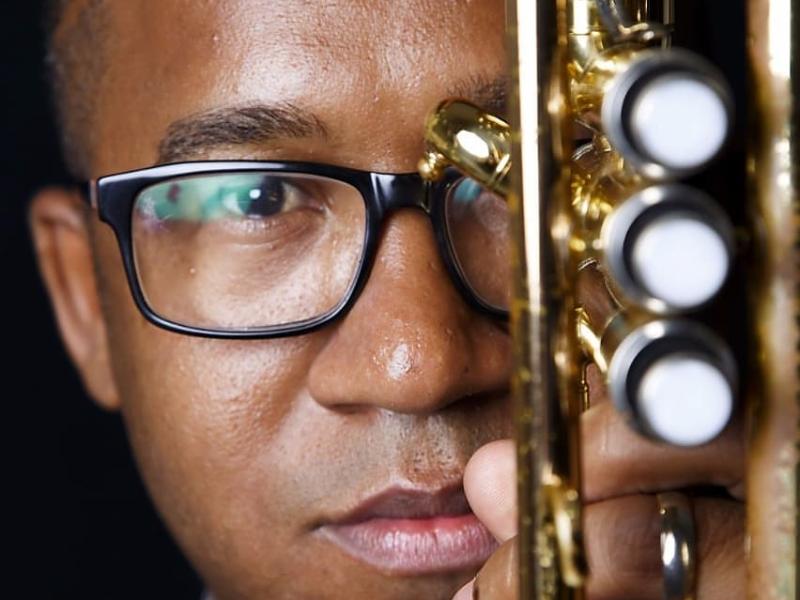 A imagem mostra um close do rosto do clarinetista Everton Machado, que usa óculos e tem expressão séria e concentrada. Ele segura um clarinete de metal dourado próximo ao rosto, com os pistões destacados em primeiro plano. A iluminação foca seu olhar e o instrumento, criando uma atmosfera de intensidade e conexão com a música. O fundo escuro realça os detalhes do instrumento e a expressão do músico.