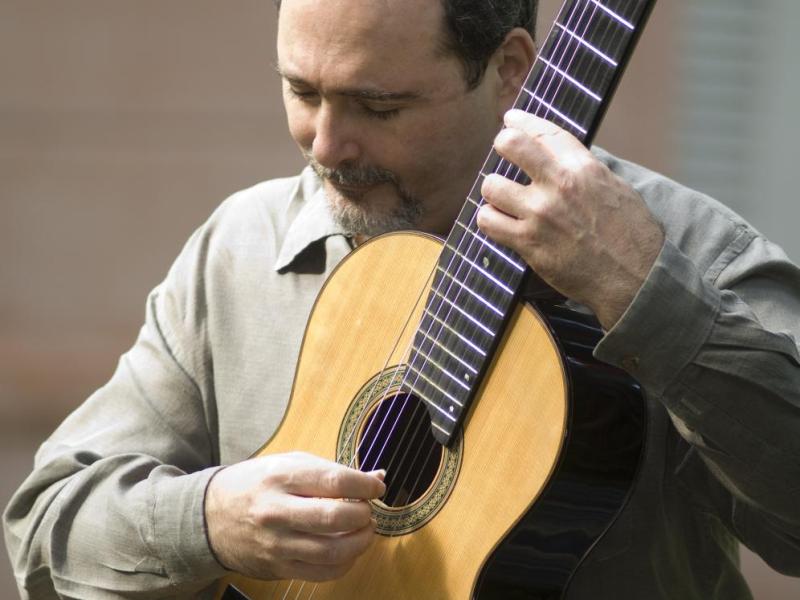A imagem mostra um homem tocando um violão clássico. Ele veste uma camisa de manga longa e parece concentrado enquanto dedilha as cordas do instrumento. O fundo desfocado e a iluminação suave realçam os detalhes do rosto do músico e do violão.