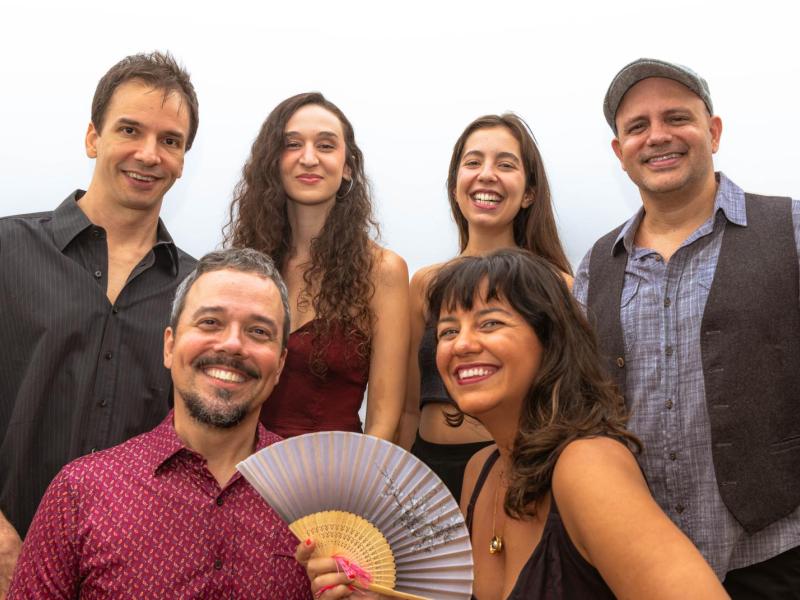 A imagem mostra os seis integrantes do grupo musical Ordinarius posando juntos e sorrindo para a câmera em um ambiente claro e descontraído. Três homens estão em pé ao fundo, enquanto duas mulheres e um homem estão à frente. Uma das mulheres segura um leque aberto. Todos demonstram alegria e sintonia, transmitindo o espírito leve, carismático e acolhedor do sexteto vocal.