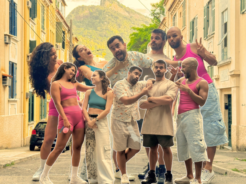 Grupo de dez pessoas posa de forma divertida em uma rua residencial com prédios coloridos e o Morro Dois Irmãos ao fundo. Três mulheres usam roupas esportivas, sendo duas em rosa e uma em azul, e sete homens usam roupas casuais ou regatas rosas. Alguns fazem poses exageradas, sorrindo, apontando ou abraçando os colegas. O clima é descontraído e alegre, com destaque para a diversidade de expressões e estilos.