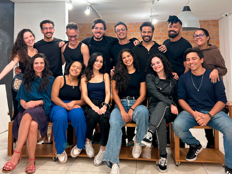 Grupo de 15 pessoas posa sorridente para a foto em ambiente interno com parede de tijolos e iluminação clara. O grupo é formado por homens e mulheres jovens, vestindo roupas casuais, majoritariamente em tons escuros. Sete pessoas estão sentadas à frente em bancos de madeira, enquanto as demais estão em pé atrás, com os braços sobre os ombros dos colegas. O clima é de amizade e descontração.