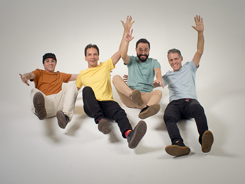 Os quatro integrantes do grupo vocal BeBossa posam sorridentes em estúdio com fundo branco, simulando estar em queda ou flutuação, com os braços erguidos e as pernas estendidas para a frente. Vestem camisetas coloridas (marrom, amarela, verde e azul) e calças casuais. A imagem transmite leveza, alegria e descontração, refletindo o espírito criativo e carismático do quarteto.