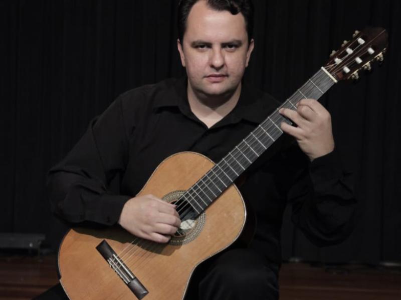 O violonista Celso Faria aparece sentado, em pose de performance, tocando um violão clássico. Ele veste camisa preta e está diante de uma cortina escura. A imagem destaca sua expressão concentrada e o instrumento de madeira com detalhes ornamentais no bojo, evidenciando o ambiente formal e dedicado à música.