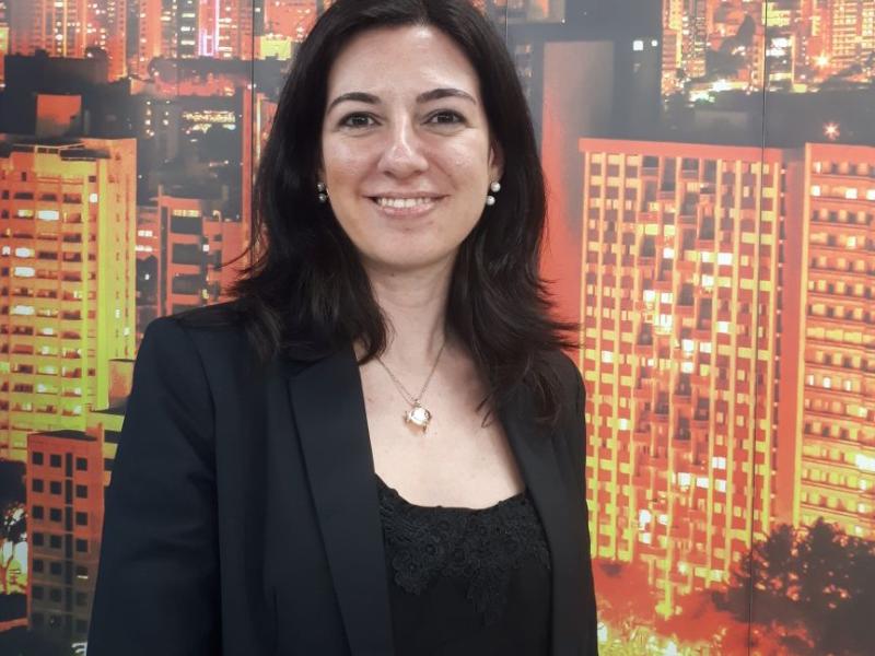 A jornalista, palestrante, escritora e consultora de comunicação Vanessa Guimarães sorri levemente diante de um painel com imagem de prédios iluminados à noite, compondo uma paisagem urbana. Ela tem cabelos escuros e lisos, na altura dos ombros, e veste blazer escuro sobre blusa preta rendada. Usa colar com pingente em formato de coração. A imagem transmite profissionalismo, simpatia e confiança.