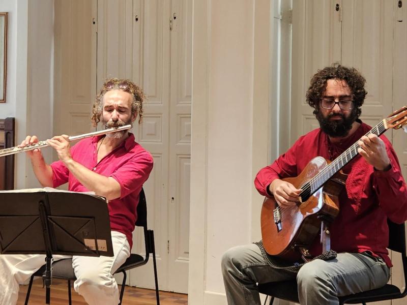 Os músicos Alexandre Bittencourt, na flauta, e Thomas Saboga, no violão, se apresentam em ambiente interno iluminado, sentados lado a lado diante de uma parede clara com portas antigas de madeira ao fundo. Ambos vestem camisas vermelhas. À esquerda, Alexandre toca flauta transversal com partitura à frente; à direita, Thomas dedilha o violão com expressão concentrada. A imagem transmite o clima intimista e refinado de uma apresentação de música de câmara.