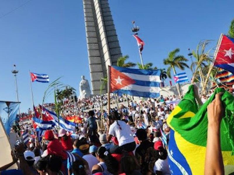 A imagem mostra uma multidão reunida em uma grande manifestação ou celebração pública em frente ao monumento a José Martí, na Praça da Revolução, em Havana, Cuba. Diversas pessoas carregam bandeiras de Cuba e também do Brasil, além de outros símbolos. O céu está limpo e azul, e palmeiras cercam o local, criando um cenário vibrante e ensolarado. A cena transmite um forte senso de união, engajamento e celebração coletiva.