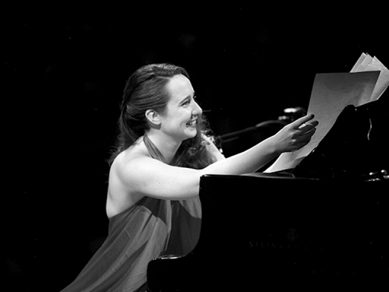 Imagem em preto e branco da cantora, compositora, pianista, atriz e neurocientista Julie Wein. Ela sorri enquanto está sentada ao piano, usando um vestido elegante. Com a mão direita, segura partituras, em um gesto que sugere preparação ou finalização de uma apresentação. Seu corpo está levemente inclinado para a frente, transmitindo leveza, carisma e envolvimento com a música.