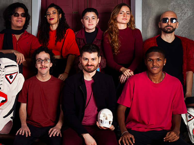 Os integrantes da banda carioca A Magnífica Máquina Maldita posam em grupo diante de uma parede cinza com portas vinho ao fundo. Quatro estão em pé na fileira de trás e quatro sentados à frente, todos vestindo roupas em tons de vermelho, bordô e preto. Um dos membros segura um crânio branco, evocando referências teatrais. Duas grandes máscaras expressivas estão apoiadas no chão à frente. A imagem transmite uma atmosfera artística, intensa e performática, característica da banda.