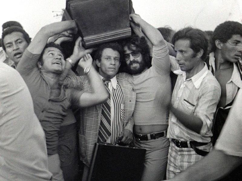 Fotografia em preto e branco de Juvenal Pereira, parte da exposição FotoRio 2025 no CCJF. A imagem retrata um momento de grande agitação popular: um homem de terno e gravata é cercado por várias pessoas, enquanto uma mala é erguida sobre sua cabeça para protegê-lo. A cena transmite a intensidade e o tumulto de um episódio coletivo marcante.