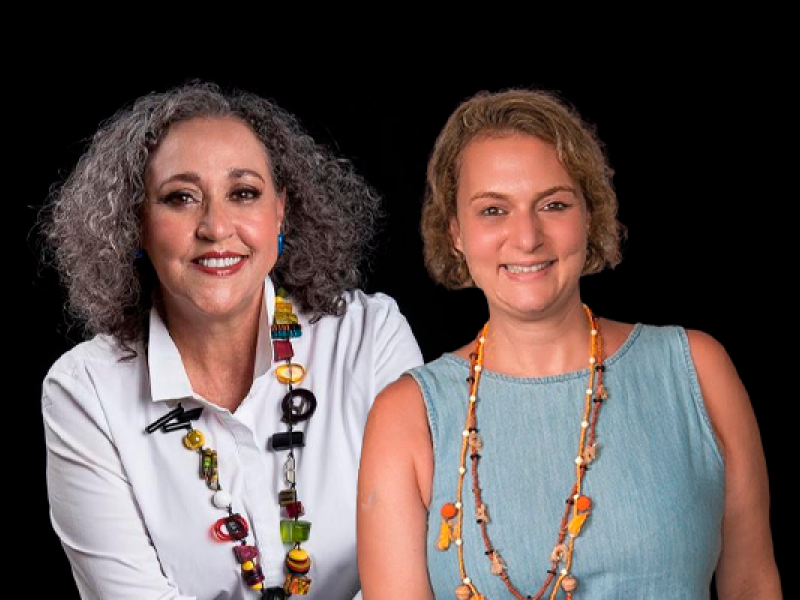 Retrato em fundo preto de duas mulheres sorrindo, lado a lado. À esquerda está Sandra Helena Pedroso, produtora e gestora cultural, com cabelos grisalhos cacheados na altura dos ombros. Ela veste camisa branca e um colar colorido com grandes contas. À direita está a jornalista Liana Varsano, de cabelos loiros curtos, usando blusa azul-clara sem mangas e colares em tons terrosos. Ambas transmitem simpatia e acolhimento.