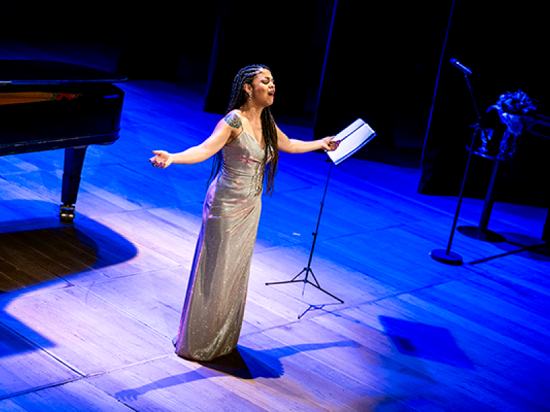 A soprano Chiara Santoro, mulher negra, apresenta-se no palco vestindo um longo vestido prateado. Ela canta de braços abertos e expressão intensa, transmitindo emoção. Ao fundo, aparecem parte de um piano de cauda e um pedestal com partitura. A cena é iluminada por luzes azuis, realçando a atmosfera solene da performance.