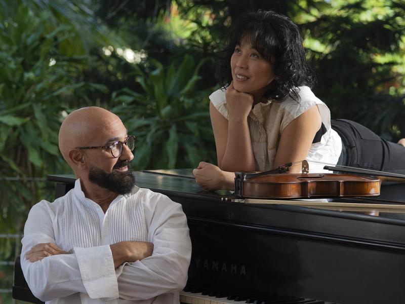 Fotografia do Duo Burajiru, formado por Fernando Thebaldi (violão) e Yuka Shimizu (piano). Em ambiente ao ar livre, Yuka está deitada sobre o piano com expressão atenta e relaxada, enquanto Fernando, sentado ao instrumento, cruza os braços e a observa com leve sorriso. Um violino repousa sobre o piano. Ao fundo, vegetação tropical completa a cena.