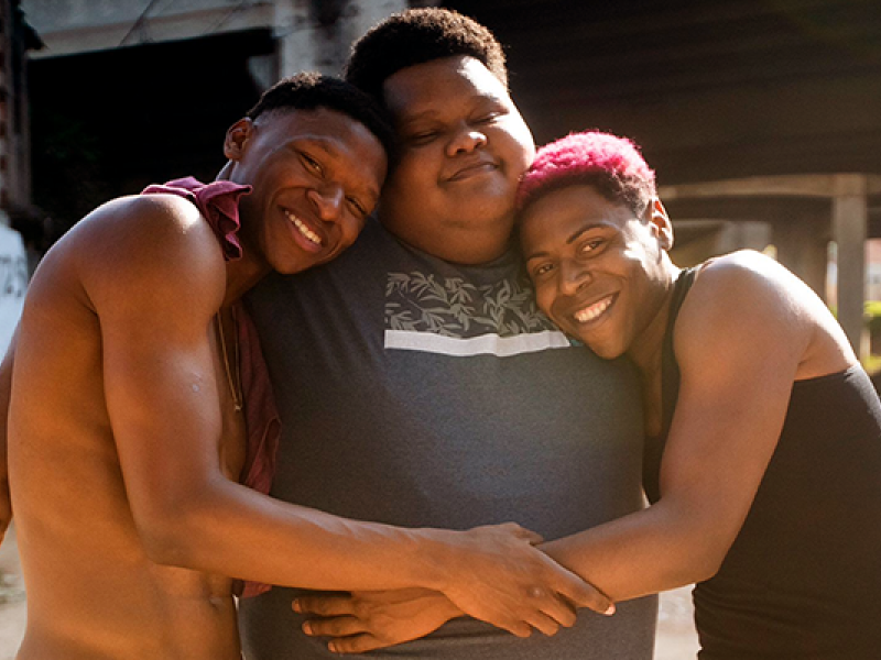 Três jovens negros posam sorridentes em um abraço coletivo, transmitindo afeto e amizade. O rapaz ao centro veste camiseta cinza estampada, enquanto os dois ao lado, um com o cabelo descolorido em tom rosado e outro com o peito nu, encostam a cabeça em seus ombros. A cena, iluminada pela luz natural, destaca a conexão e a alegria compartilhada entre eles.