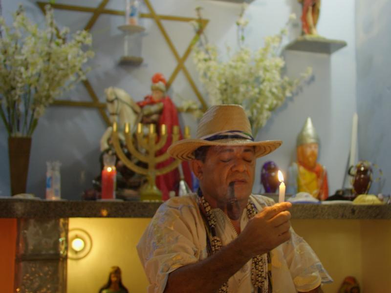 Homem com chapéu de palha e colares fuma cachimbo e aparece em frente a um altar religioso, segurando uma vela acesa com expressão de concentração e respeito. O altar é decorado com flores, imagens de santos e entidades, velas coloridas e um candelabro dourado, compondo um ambiente espiritual e ritualístico.