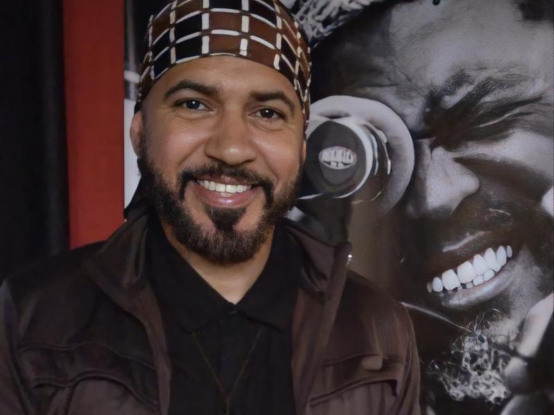 O pedagogo, curador, cineasta, roteirista e produtor Alek Lean sorri para a câmera, usando camisa preta, jaqueta marrom e um turbante estampado em marrom e branco. De barba bem aparada e expressão acolhedora, ele posa diante de um painel em preto e branco que mostra, em detalhe, o rosto de um homem sorridente, criando um cenário de forte atmosfera artística.