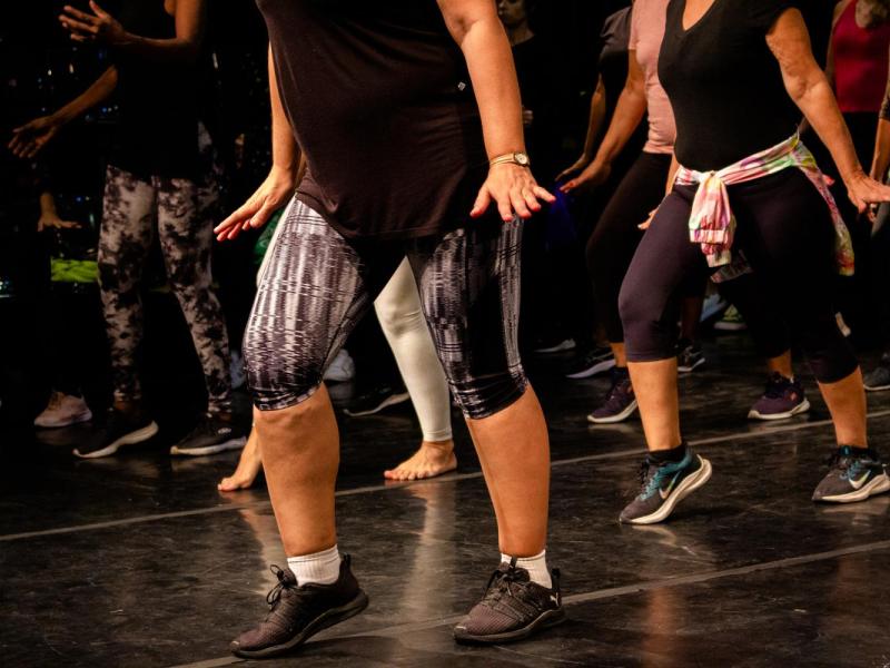 Grupo de pessoas pratica passos de dança charme em sala de ensaio. Vestindo roupas esportivas, como camisetas, leggings e tênis, algumas também estão descalças. A cena destaca o movimento das pernas em sincronia, transmitindo o ritmo marcado, o balanço característico e a energia coletiva dessa expressão cultural.