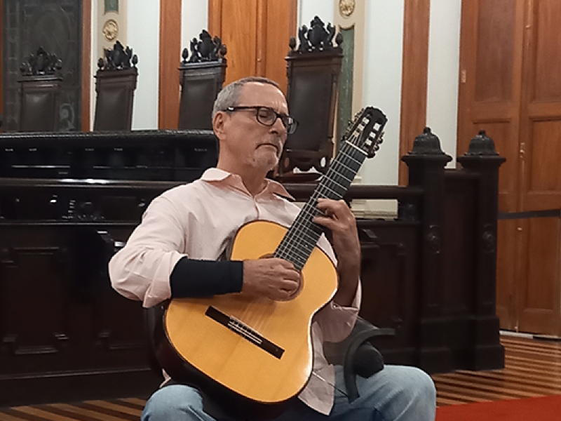 Foto da Sala de Sessões do CCJF. Ao centro, no tapete vermelho, sentado em uma cadeira, o violonista Nicolas de Souza Barros. Homem branco, cabelos grisalhos cortados rente à cabeça, óculos retangulares de hastes escuras. Ele usa uma camisa social clara, arregaçada até quase o cotovelo e calça jeans clara.. Em seu antebraço direito, ele usa uma munhequeira preta. Ele toca o violão apoiado em sua perna direita.