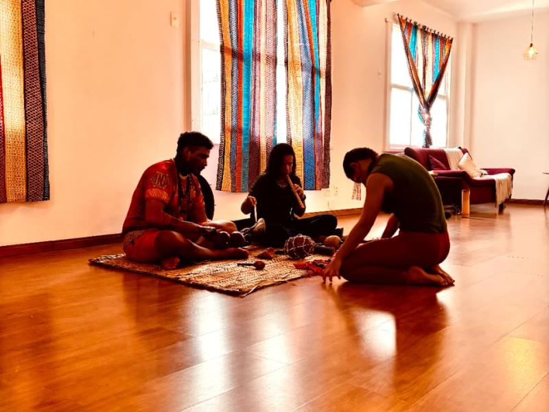 Três pessoas estão sentadas no chão de uma sala iluminada, com cortinas coloridas nas janelas ao fundo. Elas se reúnem em círculo sobre tapetes, em postura concentrada e introspectiva, sugerindo um momento de partilha, meditação ou prática ritual. O ambiente tem clima acolhedor, com sofá e almofadas ao lado, reforçando a sensação de intimidade e conexão.