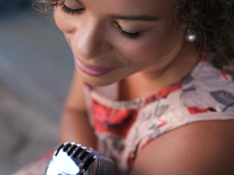A imagem mostra a cantora Debora Ondina vista de cima e de lado, com expressão serena enquanto segura um microfone vintage prateado. Morena clara, de cabelos cacheados curtos e usando um vestido estampado em tons suaves, ela transmite delicadeza e concentração, como se estivesse prestes a cantar.
